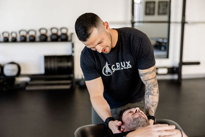 A chiropractor performs a neck adjustment on a male patient in a clinic, wearing a "Crux Sport & Spine" t-shirt. Kettlebells and gym equipment are visible in the background, highlighting the clinic's active rehabilitation focus in St. George, Utah.