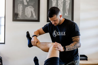  A chiropractor at Crux Sport & Spine in St. George, Utah performs a knee adjustment on a patient lying on a treatment table. The chiropractor, wearing a clinic-branded t-shirt, focuses on manipulating the patient's knee.