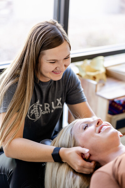 A chiropractor performs a neck adjustment on a male patient in a clinic, wearing a "Crux Sport & Spine" t-shirt. Kettlebells and gym equipment are visible in the background, highlighting the clinic's active rehabilitation focus in St. George, Utah.